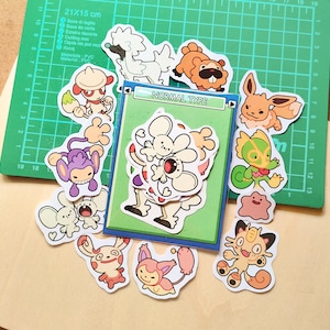 Op de afbeelding: Een verzameling kleurrijke cartoonstickers met verschillende personages, waaronder een wit hondachtig wezen, een bruin beerachtig figuur en een roze blob. Een groene snijmat met de tekst "NORMAL TYPE" op de achtergrond.