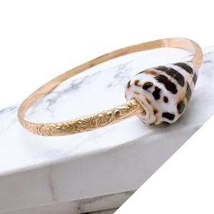 May include: A gold bangle bracelet with an intricate floral design and a large, white and brown seashell charm.