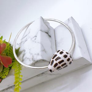 May include: A silver bracelet with a natural seashell pendant. The shell has a distinctive black and white spotted pattern. The jewellery is set against a backdrop of white marble and greenery, emphasizing the bracelet's design and texture.