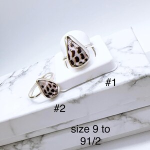Peut inclure: Deux bagues en argent avec des coquillages en forme de goutte d'eau, présentant un motif de taches marron sur fond crème. Les bagues sont exposées sur une surface blanche avec les textes "#1" et "#2". Le texte "size 9 to 9 1/2" est également visible.