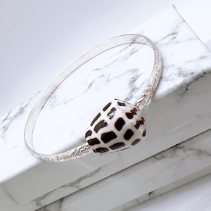 May include: A silver bangle bracelet with a natural seashell, patterned with brown and white spots. The bracelet has an etched design near the shell. The jewellery is displayed on a white marble surface.