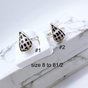 Peut inclure: Deux bagues en argent avec un motif en forme de coquillage. Les coquillages ont une base blanche avec des taches noires. Les bagues sont présentées sur une surface blanche avec le texte "#1" et "#2". Le texte "size 8 to 8 1/2" est également visible.