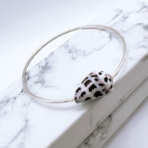 May include: A silver bangle bracelet with a natural seashell, patterned with black and white spots. The bracelet is presented on a white and grey marble surface, showcasing a minimalist design.