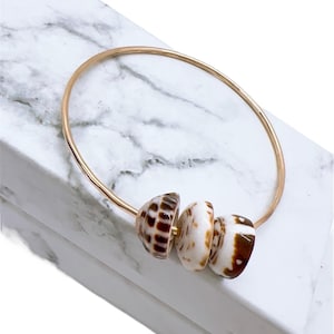 Może przedstawiać: Złota bransoletka z trzema koralikami z muszli. Koraliki są biało-brązowe, z naturalnym wzorem. Bransoletka jest prezentowana na białej marmurowej powierzchni.