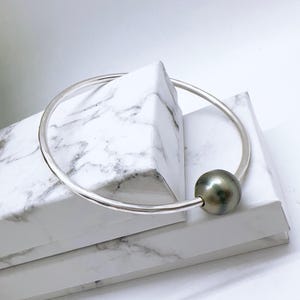 May include: A silver bangle bracelet featuring a large, dark grey pearl. The circular bracelet design has the pearl centrally positioned. The pearl has a lustrous sheen, and the bracelet is set against a white background.