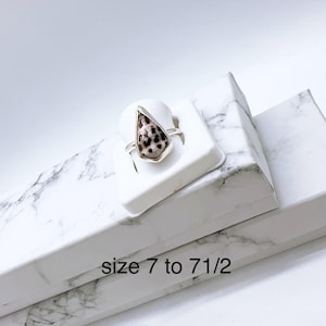 May include: A silver ring featuring a teardrop-shaped stone with a speckled pattern of brown and cream. The ring is displayed on a white stand, with the text "size 7 to 7 1/2" visible. The ring is a size 7 to 7.5.