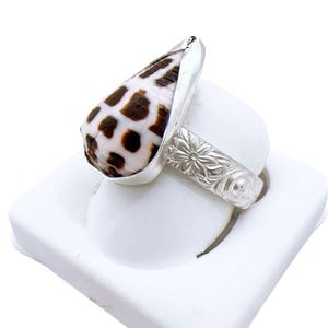 Puede incluir: Un anillo de plata con una concha en forma de cono con un patrón de manchas blancas y marrones. La banda del anillo tiene un diseño floral. El anillo se muestra sobre una superficie blanca.