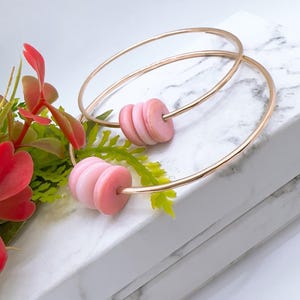 May include: A pair of gold-toned hoop earrings, each featuring three stacked, smooth, light pink circular beads. The earrings are thin, circular metal bands that hold the beads in place, creating a simple, balanced design with a polished finish.