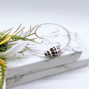 May include: A silver bracelet with a natural seashell, patterned with brown and white spots. The bracelet is displayed on a white marble surface. Greenery and flowers are visible in the background.