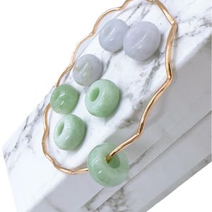 May include: A collection of jade donut-shaped beads in varying shades of green and lavender, displayed on a gold-colored, wavy metal frame. The beads rest on a white marble-patterned surface, creating a contrast of colors and textures. The arrangement suggests jewelry or decorative elements.
