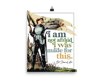 Op de afbeelding: Poster met een gestileerde illustratie van Jeanne d'Arc in harnas, met een banner. De tekst luidt: "Ik ben niet bang, ik ben hier voor gemaakt." Het kunstwerk heeft een vintage stijl met een witte rand en een zwarte clip.