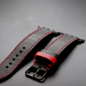 May include: A dark gray leather watch band with red stitching. The band has a black buckle and a red accent piece. The watch band is designed to fit a smartwatch. The band is made of leather.