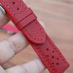 May include: Close-up of a red leather watch strap with red stitching. The strap has a textured surface and several small holes. The strap is designed to attach to a watch face.