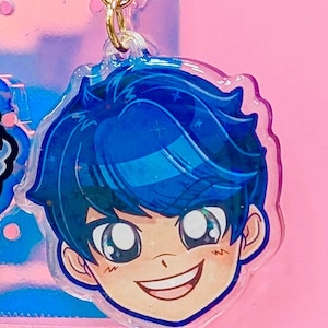 May include: A clear acrylic keychain featuring a smiling anime-style character with blue hair and eyes. The keychain has a gold-colored clasp and a pink background. The character's face is detailed with highlights and shadows.