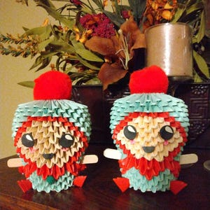 May include: Two origami figures made of paper, each with a red hat, a white face with black eyes, and a red and blue body. The figures are sitting on a wooden surface.