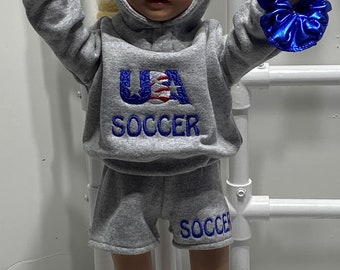 Puede incluir: Un conjunto de sudadera y pantalones cortos grises para una muñeca. La sudadera tiene un logotipo de fútbol de EE. UU. con detalles en rojo, blanco y azul. Los pantalones cortos tienen la palabra "SOCCER" bordada en la pierna. La muñeca lleva una goma de pelo azul en la muñeca.
