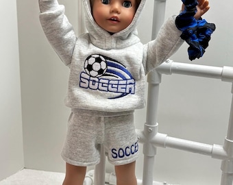 Puede incluir: Una sudadera con capucha gris y pantalones cortos a juego con la palabra "Soccer" y un gráfico de balón de fútbol. La muñeca lleva el atuendo y sostiene una diadema con estampado azul y negro.