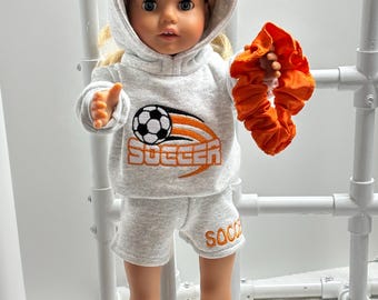 Puede incluir: Una muñeca vestida con un chándal gris con bordados de fútbol naranja. La sudadera con capucha presenta un diseño de balón de fútbol y la palabra "Soccer". La muñeca también sostiene una goma para el pelo naranja.
