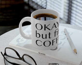 Op de afbeelding: Witte keramische koffiemok gevuld met donkere koffie, met de tekst "OKAY but first COFFEE" in zwart. De mok staat op een wit oppervlak naast een notitieblok, potlood en bril.