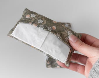 May include: A rectangular tissue holder made from fabric with a floral pattern in green, peach, and white. The holder contains white tissues. The fabric design features small flowers and leaves. The holder is being held.
