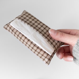 May include: A rectangular tissue holder in a brown and beige plaid design. White tissues are partially visible, emerging from the opening. The holder is made of fabric and appears soft. The item is held in a hand.