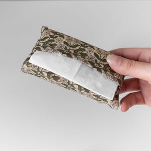 May include: A rectangular tissue holder with a floral pattern in shades of green, beige, and brown. The holder is filled with white tissues, and the design features a repeating pattern of leaves and flowers.