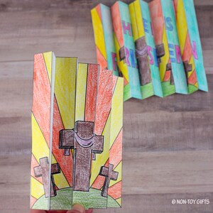 May include: A colorful paper craft with a sunrise background and three crosses. The text "He is Risen" is on the top of the craft.
