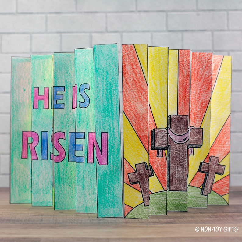 May include: A colorful paper craft with the words "He is Risen" on a green background and a sunrise scene with three brown crosses on a green hill.
