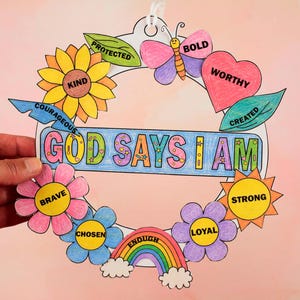 May include: A circular paper craft decoration featuring the text GOD SAYS I AM in a central blue banner. The design is surrounded by colorful hand-colored illustrations including a sunflower, butterfly, heart, rainbow, and various flowers with words like KIND, BOLD, WORTHY, PROTECTED, COURAGEOUS, BRAVE, CHOSEN, ENOUGH, STRONG, and LOYAL.