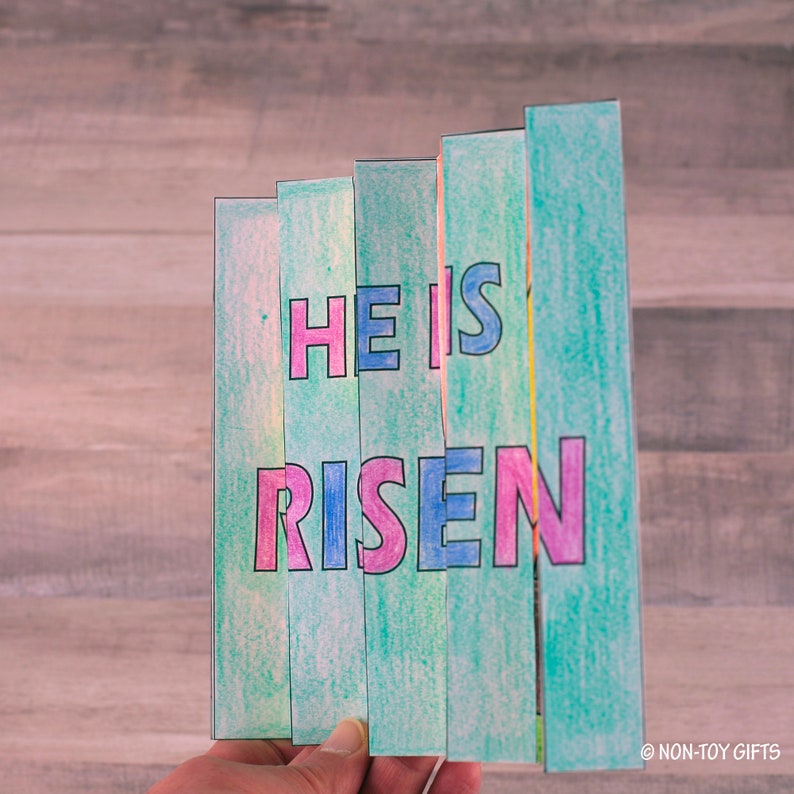 May include: A colorful paper craft with the words "He is Risen" in pink and blue letters on a green background.