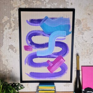 Könnte beinhalten: Abstraktes Gemälde in einem schwarzen Rahmen mit violetten, rosa und blauen Pinselstrichen auf einer beigefarbenen Leinwand. Das Gemälde hängt an einer Wand mit einem Stapel Bücher und einer Weinflasche davor.