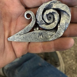 May include: A silver metal snail sculpture with a swirl design. The snail has a small tail and the letter "M" is engraved on the body.