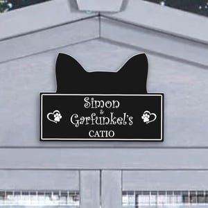 May include: A black cat-shaped sign with the words "Simon & Garfunkel's CATIO" in white lettering. The sign is mounted on a gray structure with a window and a wire mesh. The sign has paw prints and heart designs.