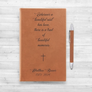 Peut inclure: Un journal en cuir marron avec une citation sur les beaux souvenirs, une croix et le nom "Matthew Brewer" avec les dates 1979-2024 gravées sur le devant. Un stylo marron est à côté du journal.