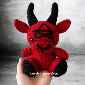 May include: A red crocheted plush toy shaped like a small devil with black horns, black ears, black feet, and a black pentagram stitched onto its forehead. The toy features two small black eyes and a black stitched mouth. Text reads LovelyTreasureNest.