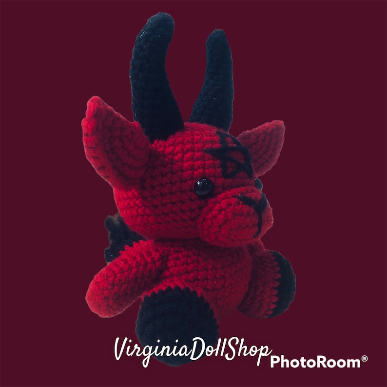 May include: A red and black crocheted plush doll featuring two prominent black horns, pointed ears, and a black pentagram symbol stitched onto its forehead. The doll has black plastic eyes, a black stitched nose, and black accents on its feet.