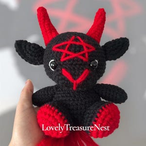 May include: A black crocheted plush toy featuring two red horns, a red pentagram stitched on the forehead, and red accents on the feet. The creature has black ears, black glossy eyes, and a red stitched nose. The text LovelyTreasureNest is visible.