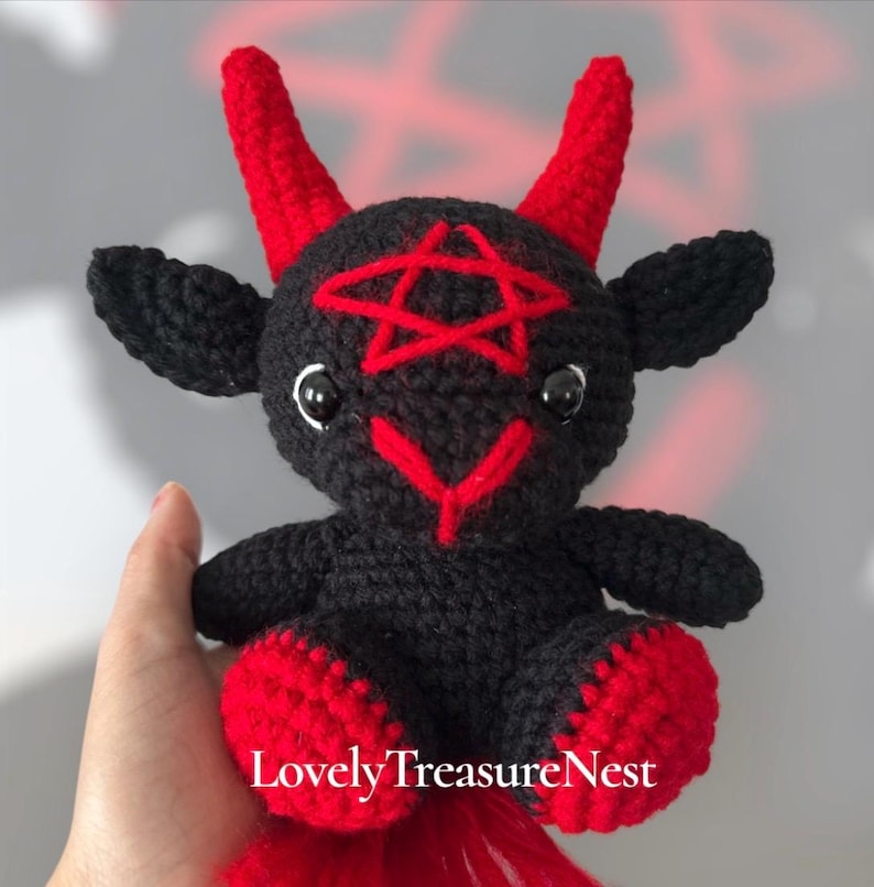 May include: A black crocheted plush toy featuring two red horns, a red pentagram stitched on the forehead, and red accents on the feet. The creature has black ears, black glossy eyes, and a red stitched nose. The text LovelyTreasureNest is visible.