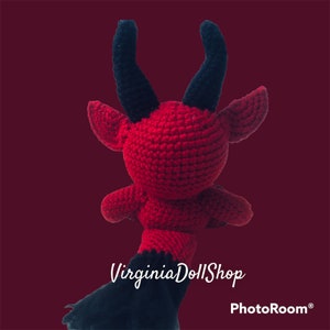 May include: A red crocheted doll featuring two prominent black horns, pointed ears, and a black lower section. The text VirginiaDollShop is printed in white across the center of the doll, with the word PhotoRoom appearing in the bottom right corner.