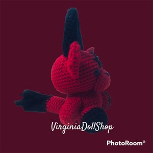 May include: A handmade crochet doll featuring a vibrant red body with contrasting black accents on the ears, feet, and tail. The creature has a rounded head, small dark eyes, and a textured yarn surface. The text VirginiaDollShop is visible.