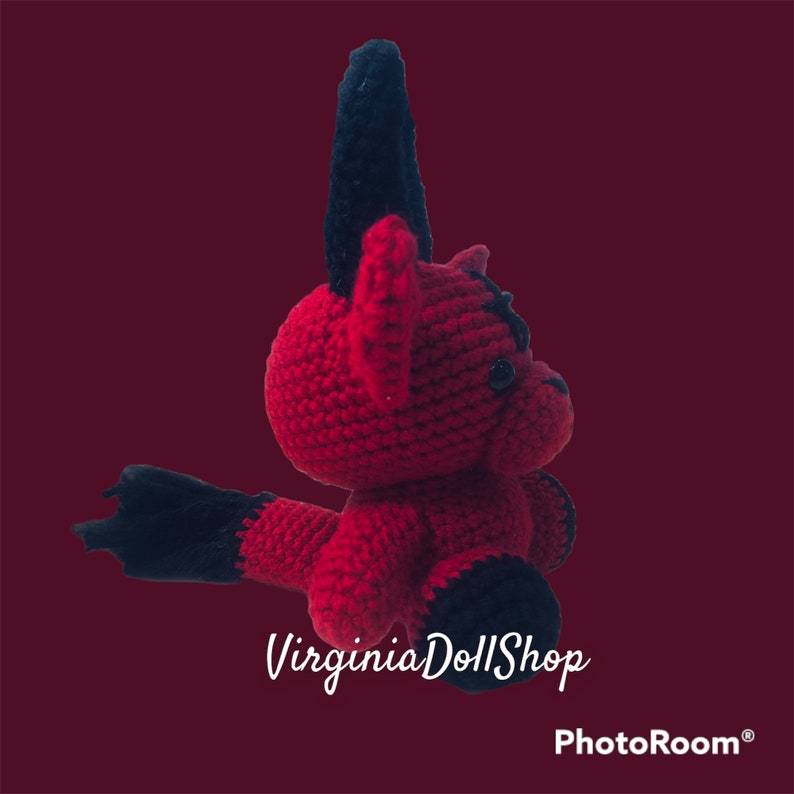May include: A handmade crochet doll featuring a vibrant red body with contrasting black accents on the ears, feet, and tail. The creature has a rounded head, small dark eyes, and a textured yarn surface. The text VirginiaDollShop is visible.
