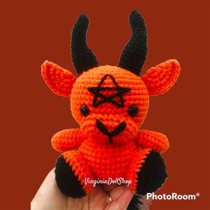May include: A bright orange crocheted plush toy shaped like a goat with two large black horns, black ears, and black hooves. A black pentagram is stitched onto the forehead, and the face features two small black bead eyes and a black stitched nose.