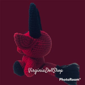 May include: A small, handmade crochet plush toy featuring a vibrant red body with contrasting black horns, black feet, and a black-tipped tail. The creature has a rounded head and pointed ears, crafted with visible yarn stitches throughout the design.