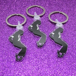 May include: Three black keychains with a silhouette of a person in a dance pose. Each keychain has a silver-colored ring and a clear plastic attachment. The keychains are on a sparkly purple background.