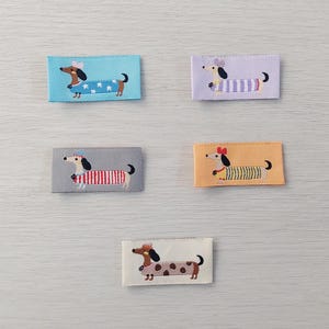 May include: Five embroidered patches featuring dachshund dogs in various outfits. The patches are rectangular with backgrounds in blue, lavender, gray, orange, and cream. The dogs wear sweaters, hats, and bows, with different patterns like stars, stripes, and polka dots.