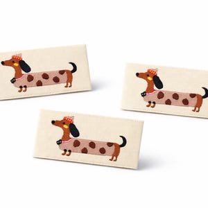Könnte beinhalten: Drei rechteckige Aufnäher mit gestickten Dackel-Designs. Die Hunde haben braune Körper mit beigefarbenen Flecken, schwarze Ohren und tragen Hüte. Die Aufnäher befinden sich auf weißem Hintergrund.