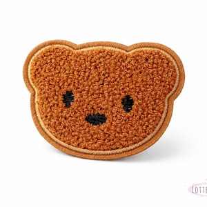 Könnte beinhalten: Brauner Teddybär-Aufnäher mit strukturiertem, plüschigem Aussehen. Das Gesicht des Bären hat schwarz gestickte Augen und Nase mit einem hellbraunen Rand. Der Aufnäher ist wahrscheinlich für Kleidung oder Accessoires.