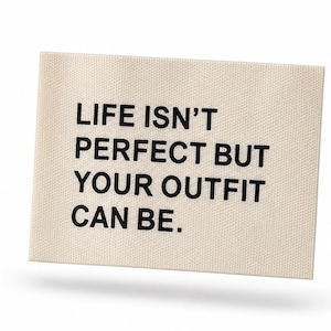 Könnte beinhalten: Ein rechteckiges, cremefarbenes Textiletikett mit dem in Schwarz aufgedruckten Text LIFE ISN'T PERFECT BUT YOUR OUTFIT CAN BE. Das Etikett hat eine gewebte Struktur und klare, gerade Kanten, die eine gut lesbare Botschaft in serifenloser Schrift zeigen.
