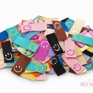 Könnte beinhalten: Ein Haufen bunter Stoffetiketten mit gestickten Smiley-Gesichtern. Die rechteckigen Etiketten sind in verschiedenen Farben erhältlich, darunter Rosa, Blau, Schwarz und Braun. Die Smiley-Gesichter sind weiß.