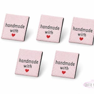 Könnte beinhalten: Fünf rosa Stoffetiketten mit dem Text "handmade with" und einem roten Herz. Die Etiketten haben einen schwarzen Rand und sind auf weißem Hintergrund angeordnet. Für handgefertigte Artikel.